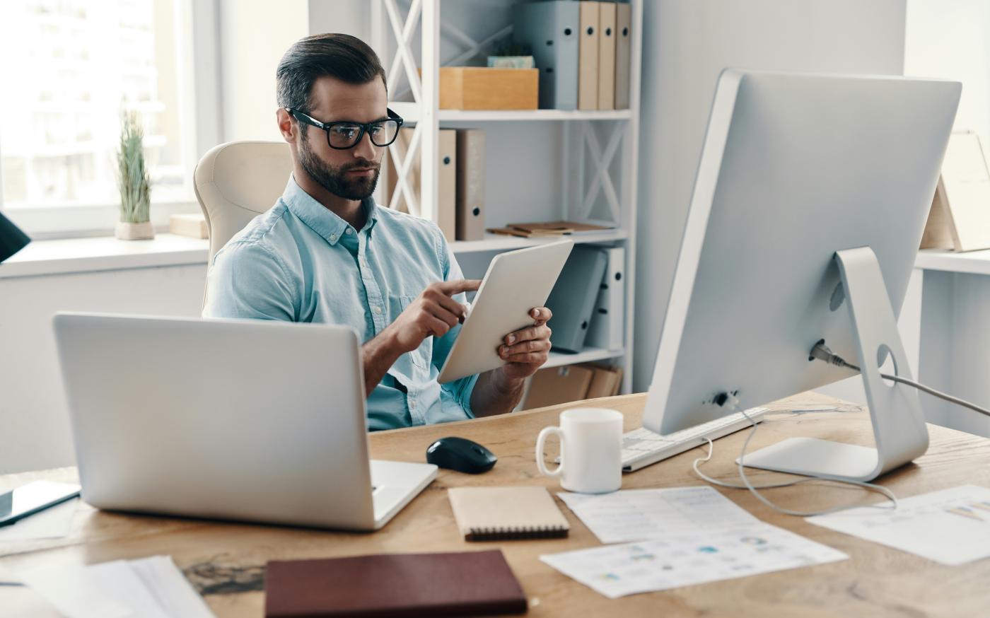 A businessman sitting at an office desk working on a tablet, with a desktop computer, coffee mug, and notebooks nearby.