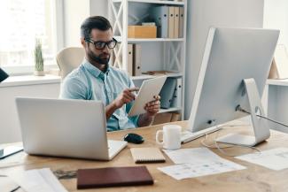 A businessman sitting at an office desk working on a tablet, with a desktop computer, coffee mug, and notebooks nearby.
